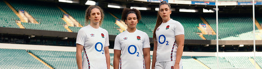 Three women in white English rugby jerseys pose in a stadium during Castore Umbro licensing announcement.