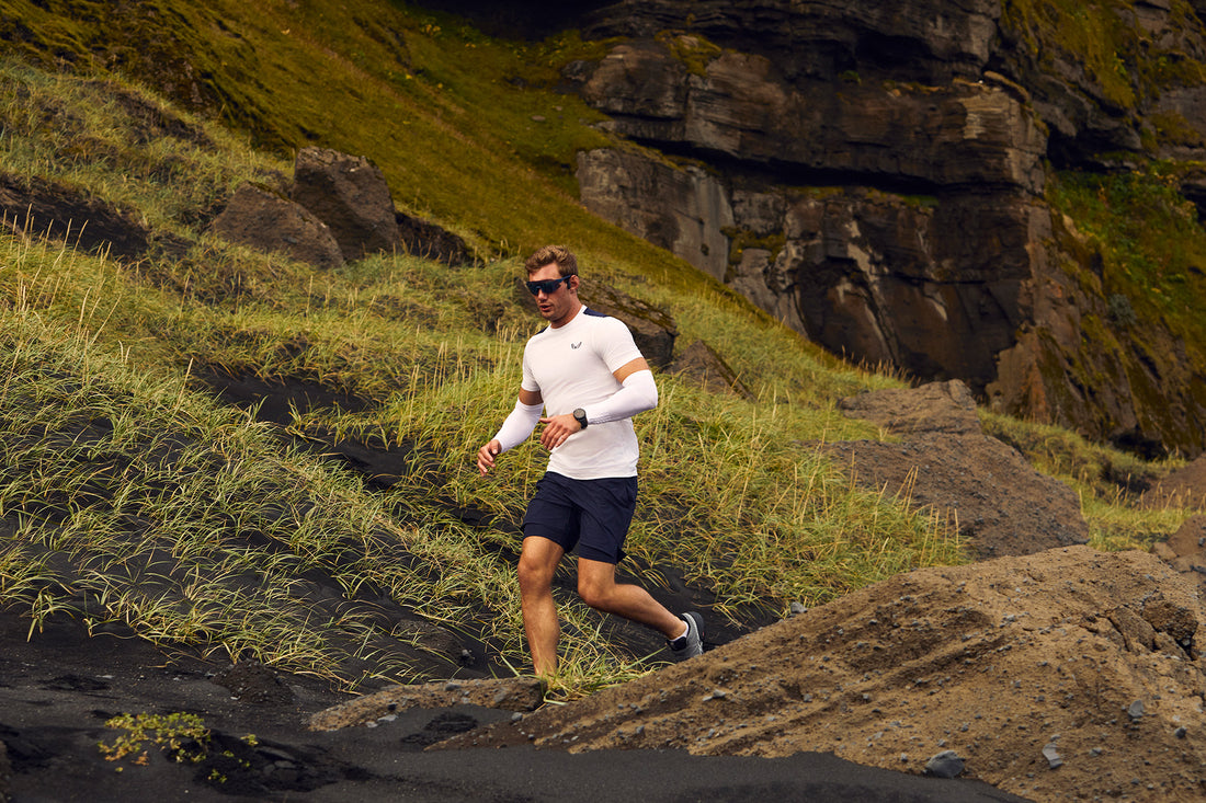 Male runner in a white long sleeve top and black shorts running along a rugged rocky trail with grassy slopes.