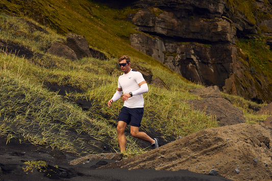 Male runner in a white long sleeve top and black shorts running along a rugged rocky trail with grassy slopes.