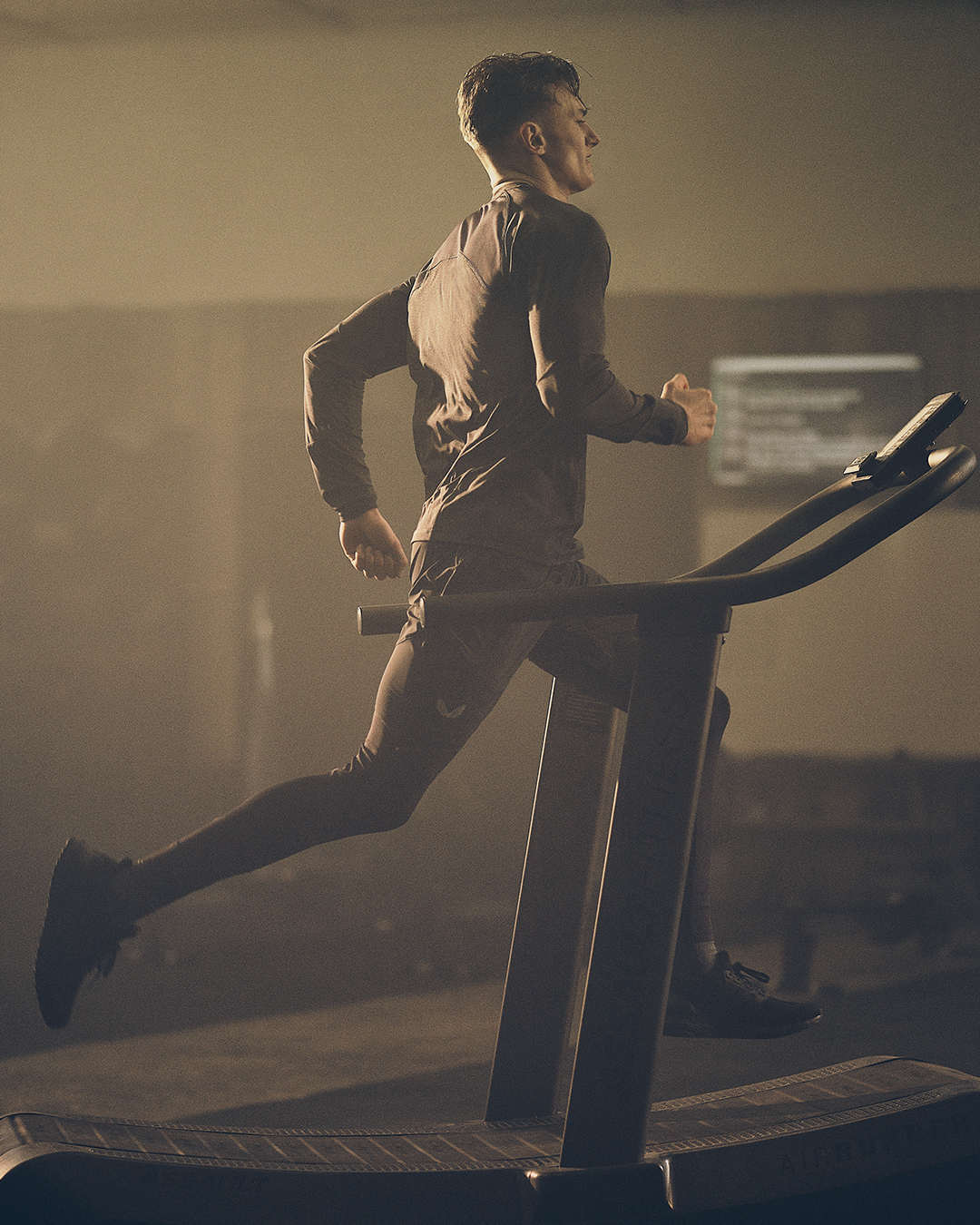 Man running on a treadmill in a dim gym wearing a dark performance top