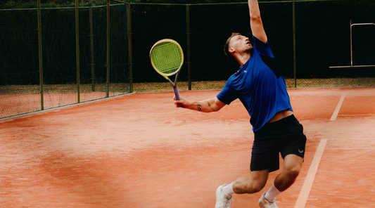 Tennis player in a blue shirt serves on a red clay court with a green racket.