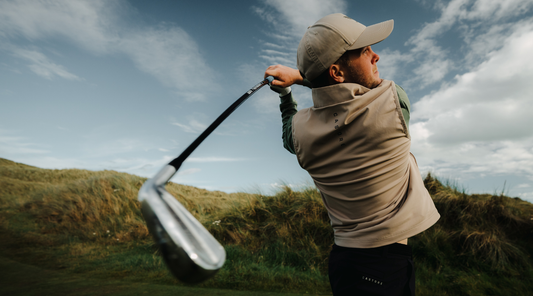 Golfer wearing a beige cap and vest swings a golf club on a sunny coastal course.