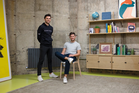 Two men in Castore branded apparel pose in a modern office with a concrete wall and wooden shelves