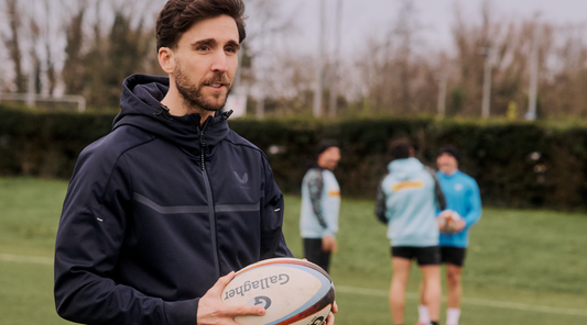 Man in a dark jacket holding a Gilbert rugby ball during a training session with teammates in the background