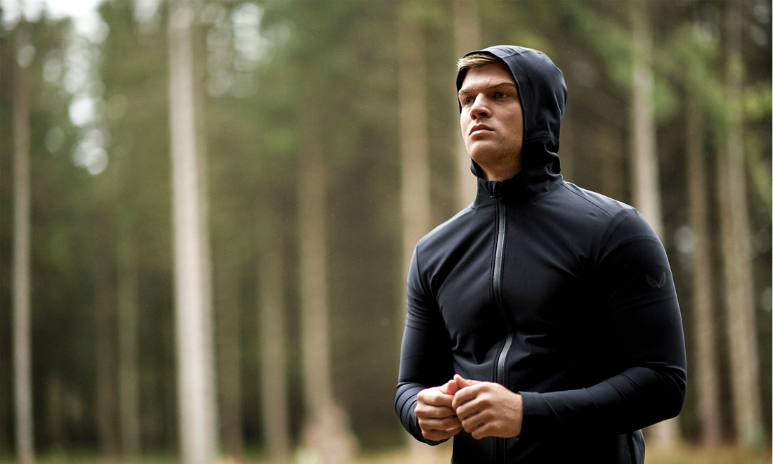 Man in a black hooded winter jacket stands in a forest during an outdoor workout.
