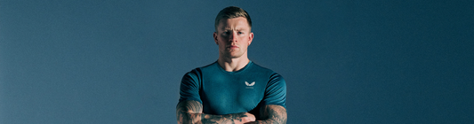 Athlete with tattoos wearing a teal athletic shirt stands against a blue backdrop.