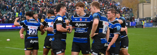 Bath Rugby players in blue and black striped home kit celebrate on the field with fans in the stands