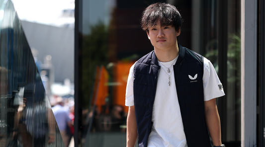 Young man in a white tee and dark vest walking beside a glass storefront in a sunny urban setting