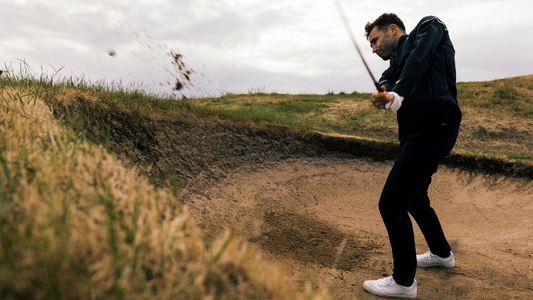 Golfer in a dark jacket and white sneakers hits a bunker shot from a sandy trap as sand sprays into the air.
