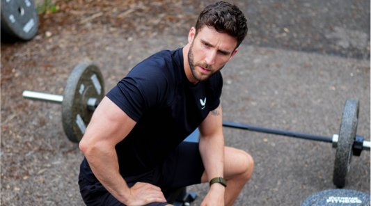 Fit man in a black athletic shirt kneels beside a barbell with weights outdoors during a workout