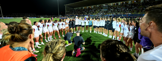 Women rugby players form a close circle huddle on the lit field after a game.