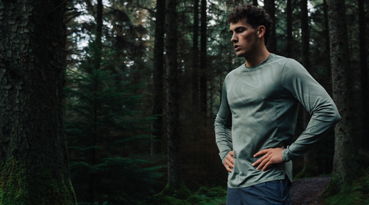 Man in a light green patterned athletic top stands with hands on hips in a dense forest.