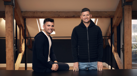 Two men in a bright indoor studio with exposed wooden beams, one seated at a table and the other standing in a black quilted vest.