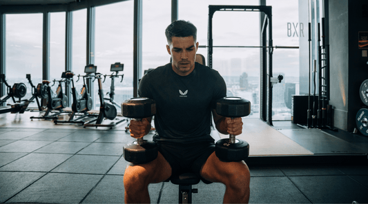 Athlete in a black Castore shirt seated in a gym performs a heavy dumbbell curl with gym equipment in the background