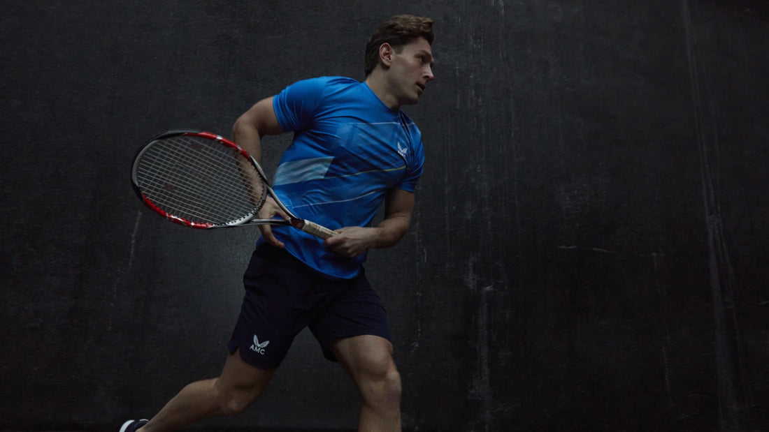 Male tennis player in a blue athletic shirt and dark shorts with a racket on a dark court backdrop.