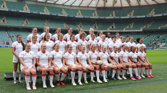 Group of female rugby players in white kits pose for a team photo on a stadium field.