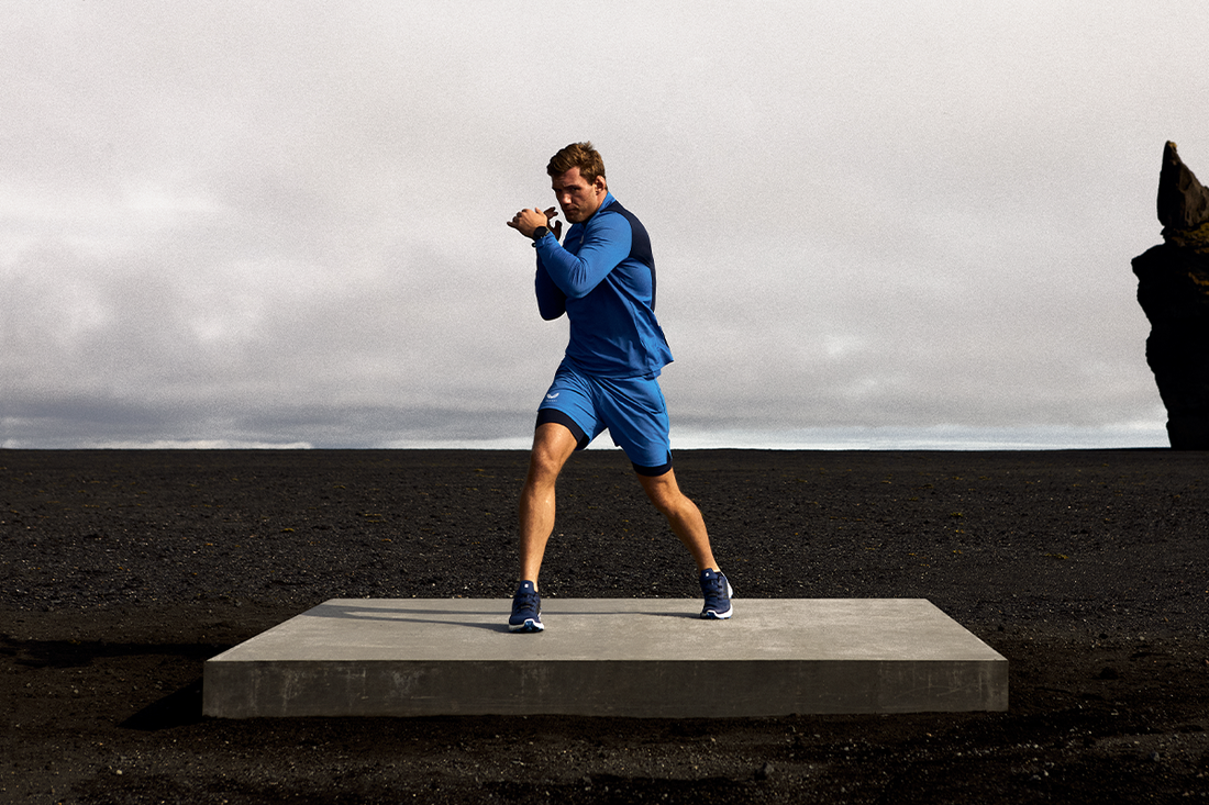 Male athlete in blue training wear mid stride on a dark volcanic field beneath a cloudy sky with a rocky outcrop.