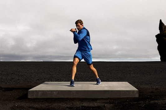 Male athlete in blue training wear mid stride on a dark volcanic field beneath a cloudy sky with a rocky outcrop.