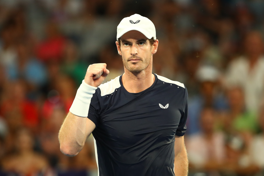 Andy Murray on a tennis court, fist raised in navy Castore kit and white cap during a press feature.