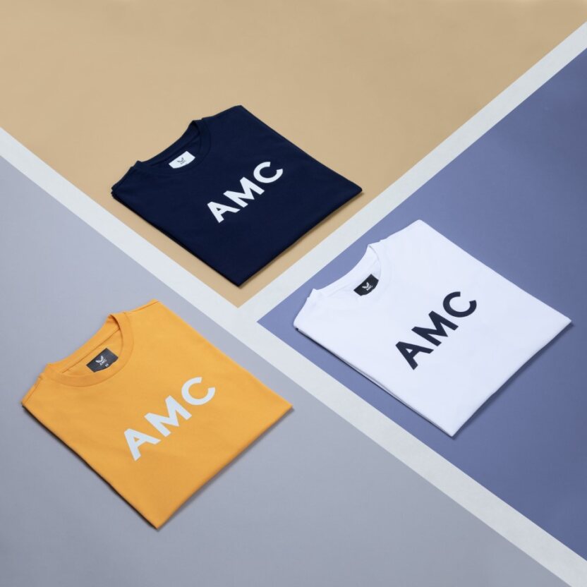 Flat lay of four AMC logo T shirts in navy orange and white on a geometric backdrop