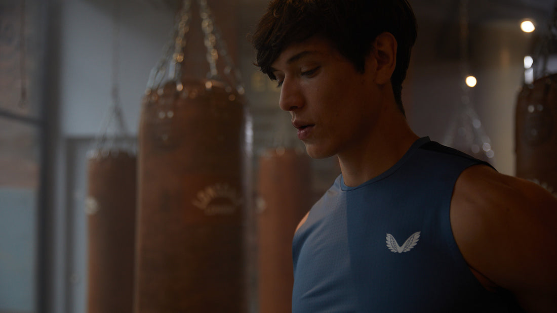 Side profile of a young man in a blue sleeveless top in a gym with hanging punching bags