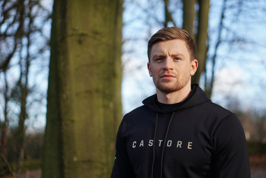 Man in a black Castore hoodie posing outdoors among trees in a forest.