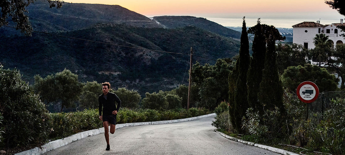 Runner in black athletic gear on a winding road at sunset with hills and shrubs in the background.