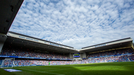 Wide view of a Rangers FC stadium with blue seating and a cloudy sky