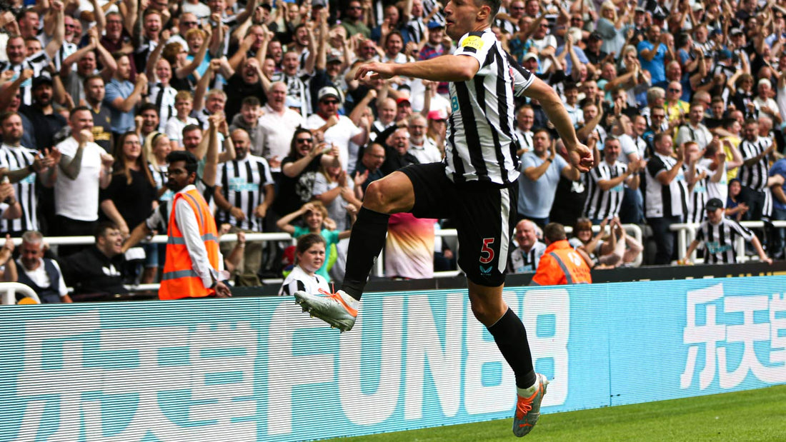 Footballer in black and white striped kit leaps near the sideline as fans celebrate in the stands