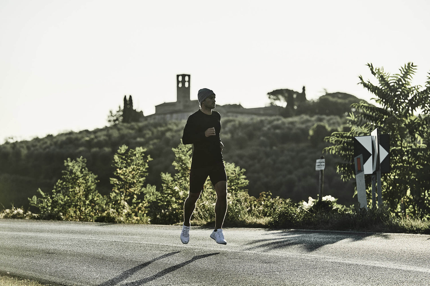 Runner in dark athletic wear on a sunlit rural road with hills and a distant church in the background.