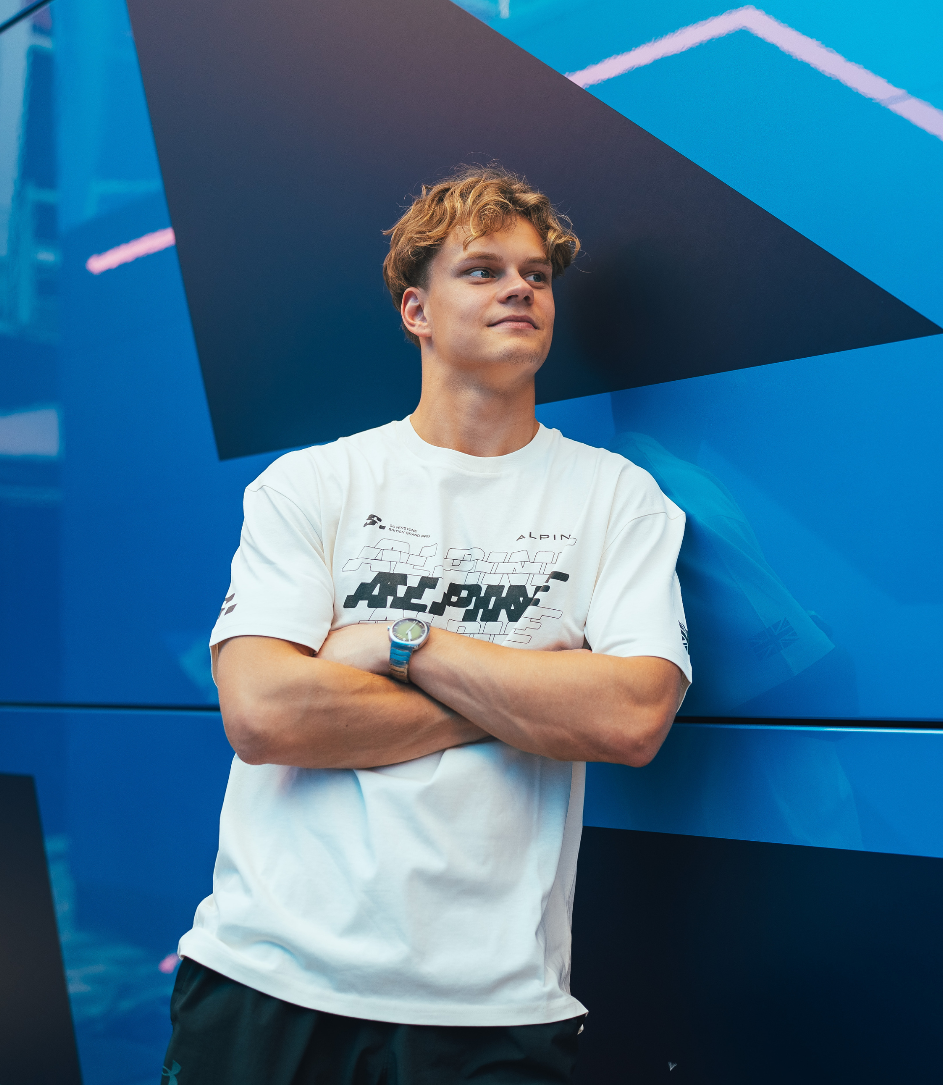 Young man wearing a white oversized Alpine T shirt with bold graphic print against a blue geometric wall.