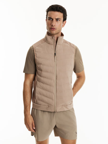 A man wearing a light beige zip-up puffer vest over a matching beige short-sleeve athletic shirt and shorts, photographed against a plain white studio background.