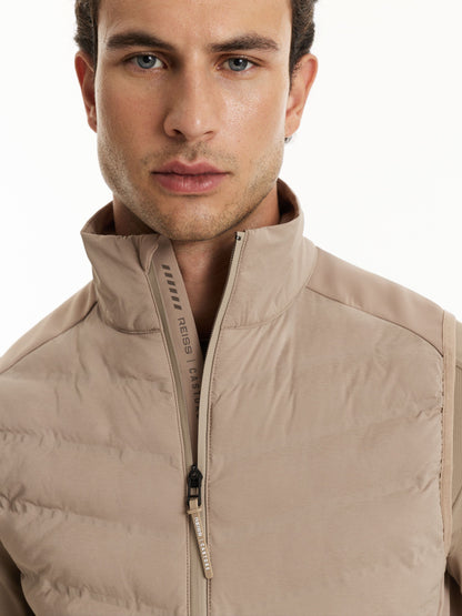 A close-up shot of a man wearing a beige puffer vest with a high stand-up collar and a front zipper. The brand name 'reaps' is visible on the zipper pull tab.