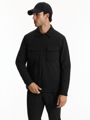 A man wearing a black zip-up jacket with two chest flap pockets, black pants, and a black baseball cap, photographed against a plain white studio background.