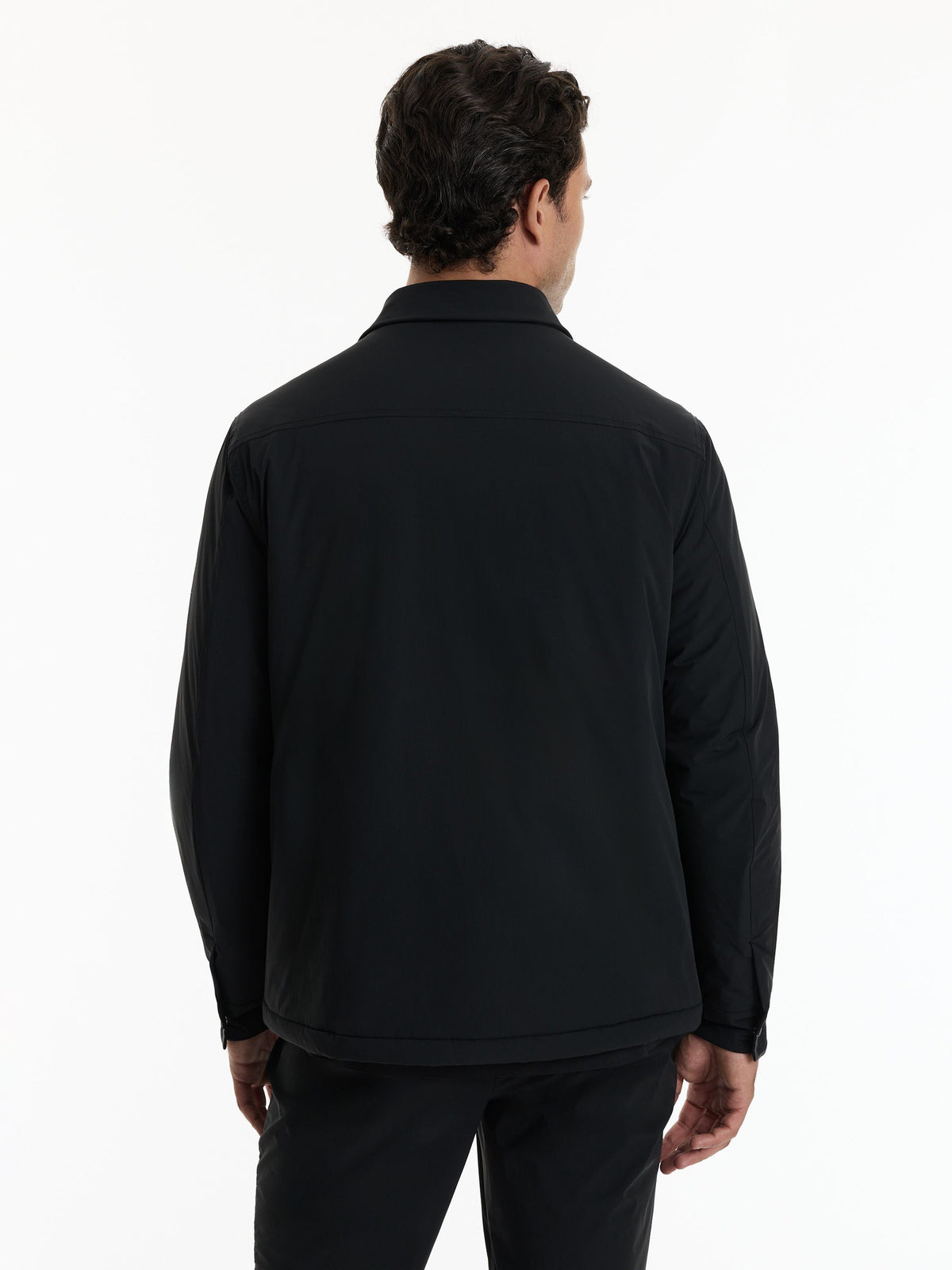 A man viewed from the back wearing a black long-sleeve collared jacket and black trousers against a plain white studio background.