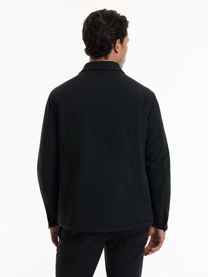 A man viewed from the back wearing a black long-sleeve collared jacket and black trousers against a plain white studio background.