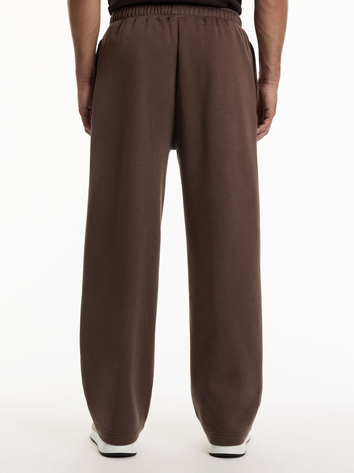 A person viewed from the back wearing loose, wide-leg brown sweatpants with an elastic waistband, shown against a plain white studio background.