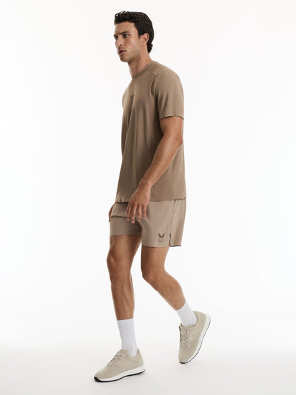A man in a matching taupe brown athletic set consisting of a short-sleeve t-shirt and shorts, paired with white socks and beige sneakers, walking against a plain white studio background.