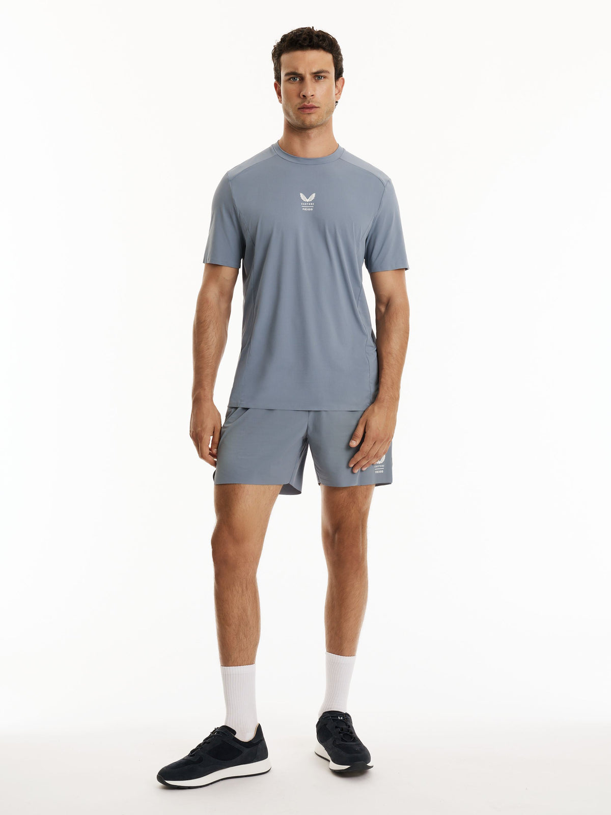 A man modeling a light blue-gray athletic t-shirt and matching shorts set, featuring a small white logo on the chest, paired with white socks and dark sneakers, against a plain white studio background.