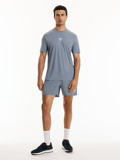 A man modeling a light blue-gray athletic t-shirt and matching shorts set, featuring a small white logo on the chest, paired with white socks and dark sneakers, against a plain white studio background.