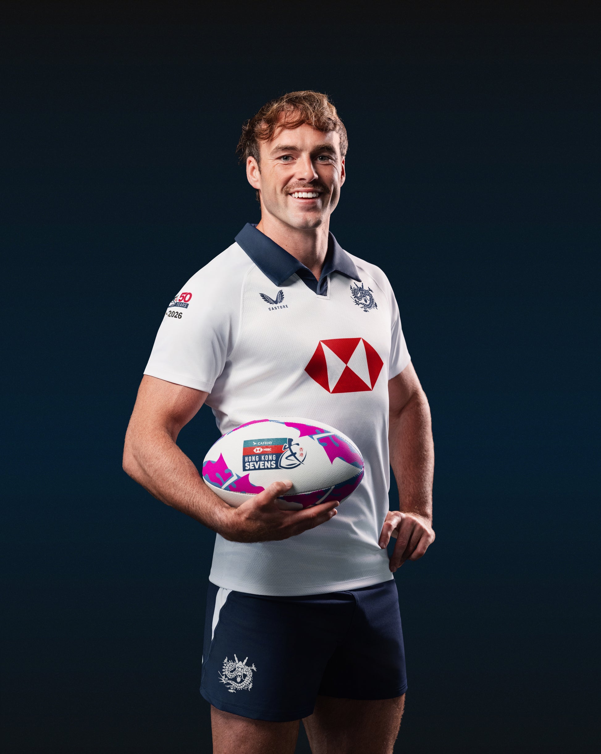 Male model wearing a white Hong Kong China Rugby away jersey with navy polo collar and gold dragon crest.