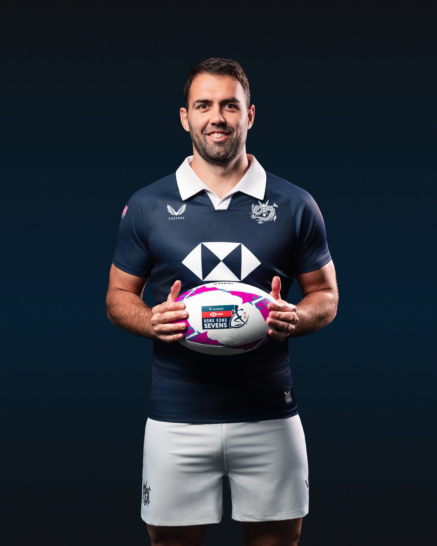 Athlete in navy HK7s jersey with white collar and crest, white shorts, holding a rugby ball against a dark backdrop