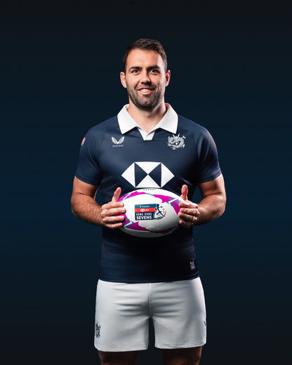Athlete in navy HK7s jersey with white collar and crest, white shorts, holding a rugby ball against a dark backdrop