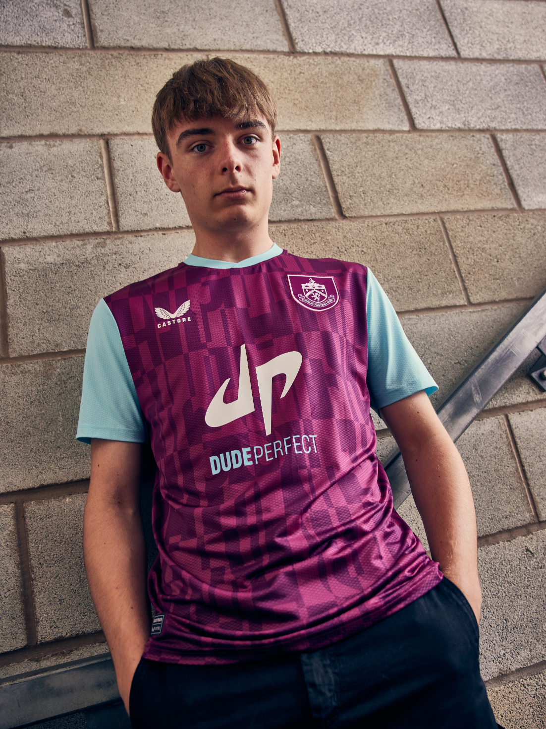 Burnley FC 24/25 junior home shirt in maroon with a purple pattern and aqua sleeves, featuring the club crest and Dude Perfect sponsor.