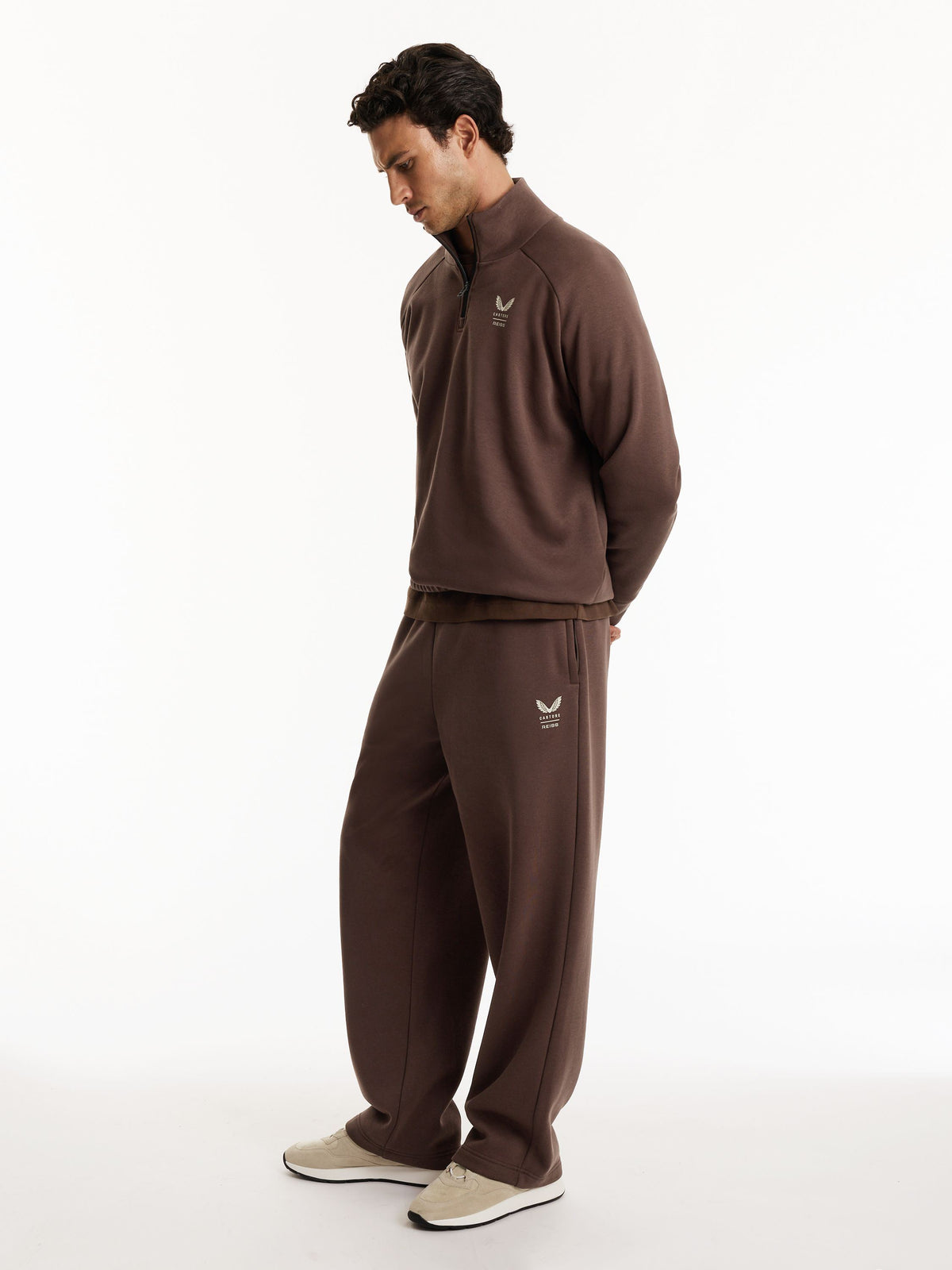 A man wearing a matching brown tracksuit, consisting of a quarter-zip pullover and matching sweatpants, standing against a plain white studio background.