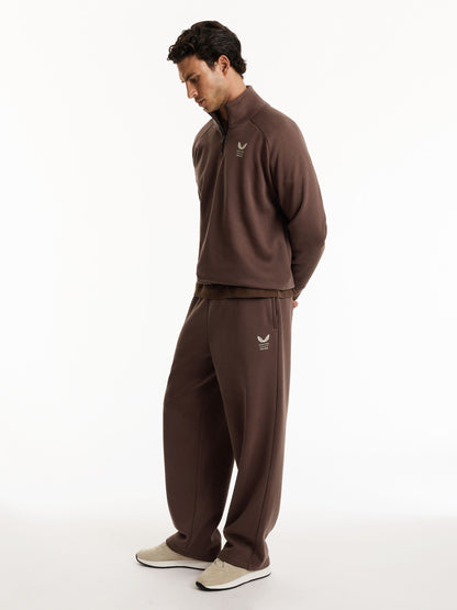 A man wearing a matching brown tracksuit, consisting of a quarter-zip pullover and matching sweatpants, standing against a plain white studio background.