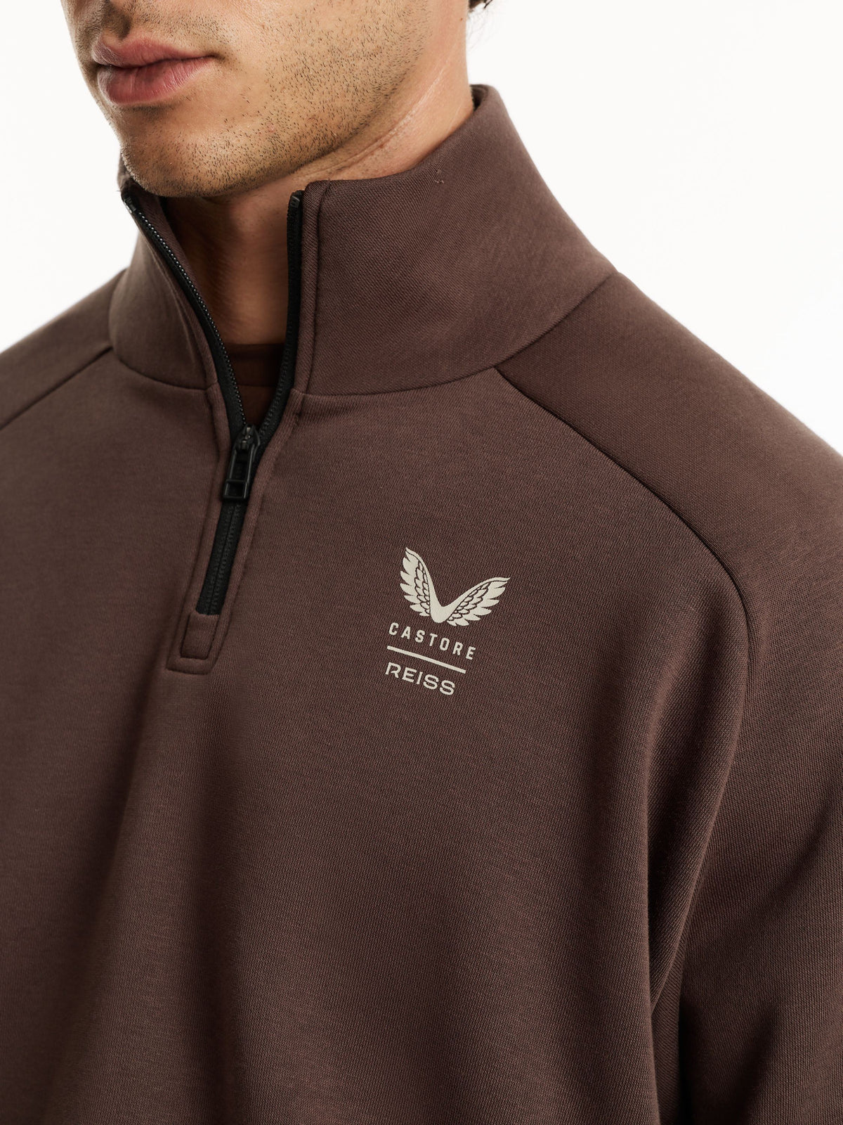 A close-up of a man wearing a brown quarter-zip pullover sweatshirt with a white embroidered winged logo and the text 'CASTORE REISS' on the left chest.