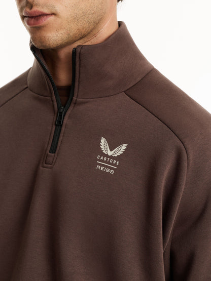 A close-up of a man wearing a brown quarter-zip pullover sweatshirt with a white embroidered winged logo and the text 'CASTORE REISS' on the left chest.