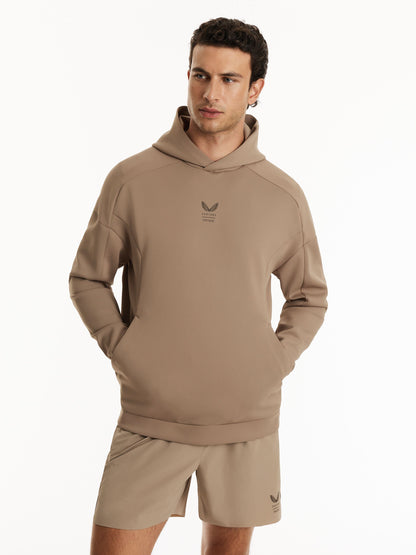 A man wearing a taupe-brown Adidas hoodie and matching shorts, standing against a plain white studio background.