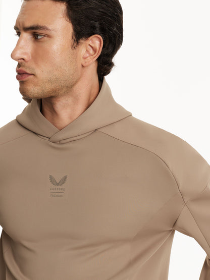 A close-up of a man wearing a beige hooded long-sleeve athletic shirt with a small winged logo and the text 'CASTORE' and 'REISS' on the left chest, against a clean white background.